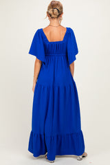 woman wearing a bright blue maternity maxi dress with flutter sleeves and a tiered skirt, showing the back view