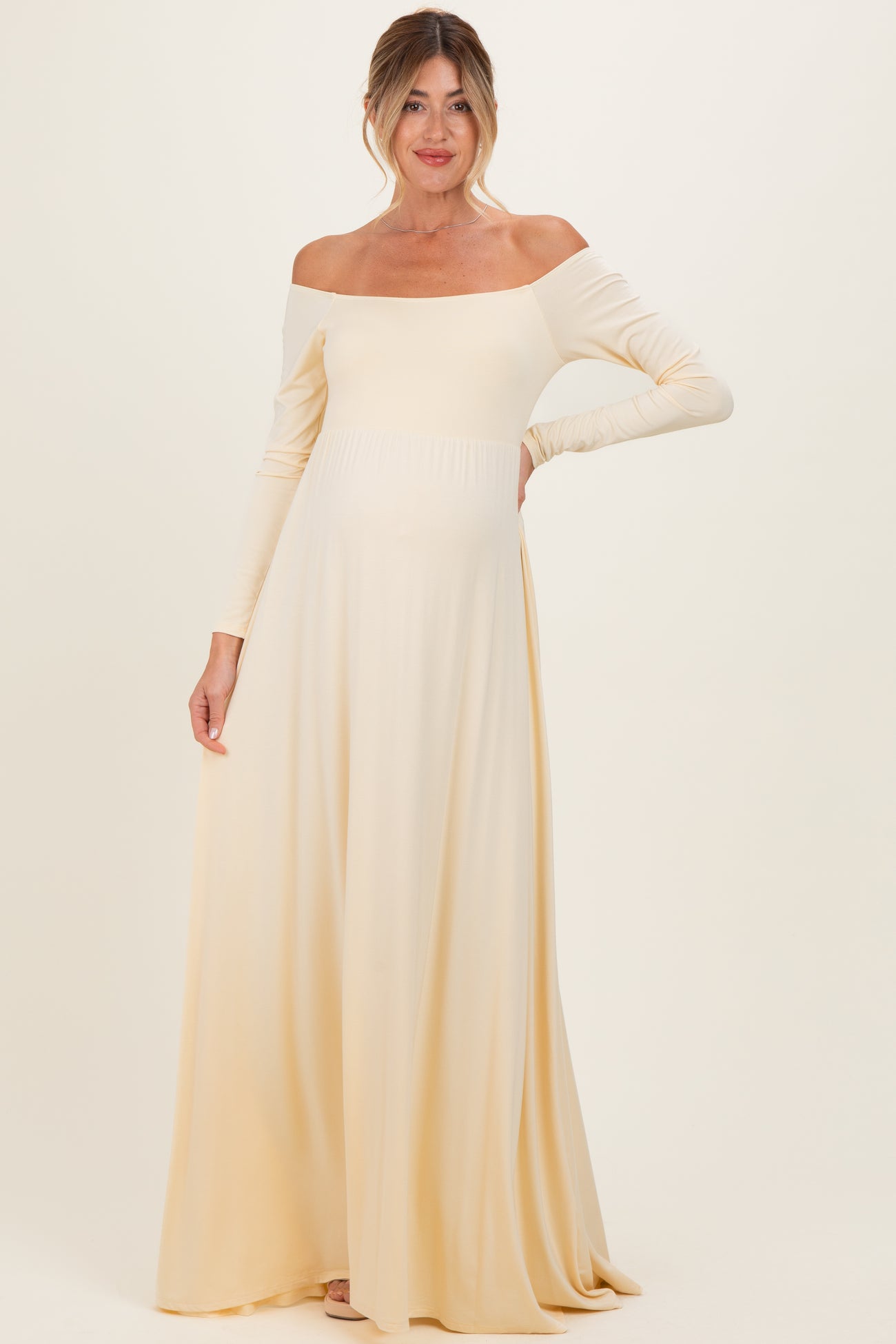 woman wearing a pale yellow off-the-shoulder maternity gown with a flowing skirt and long sleeves, showcasing her baby bump