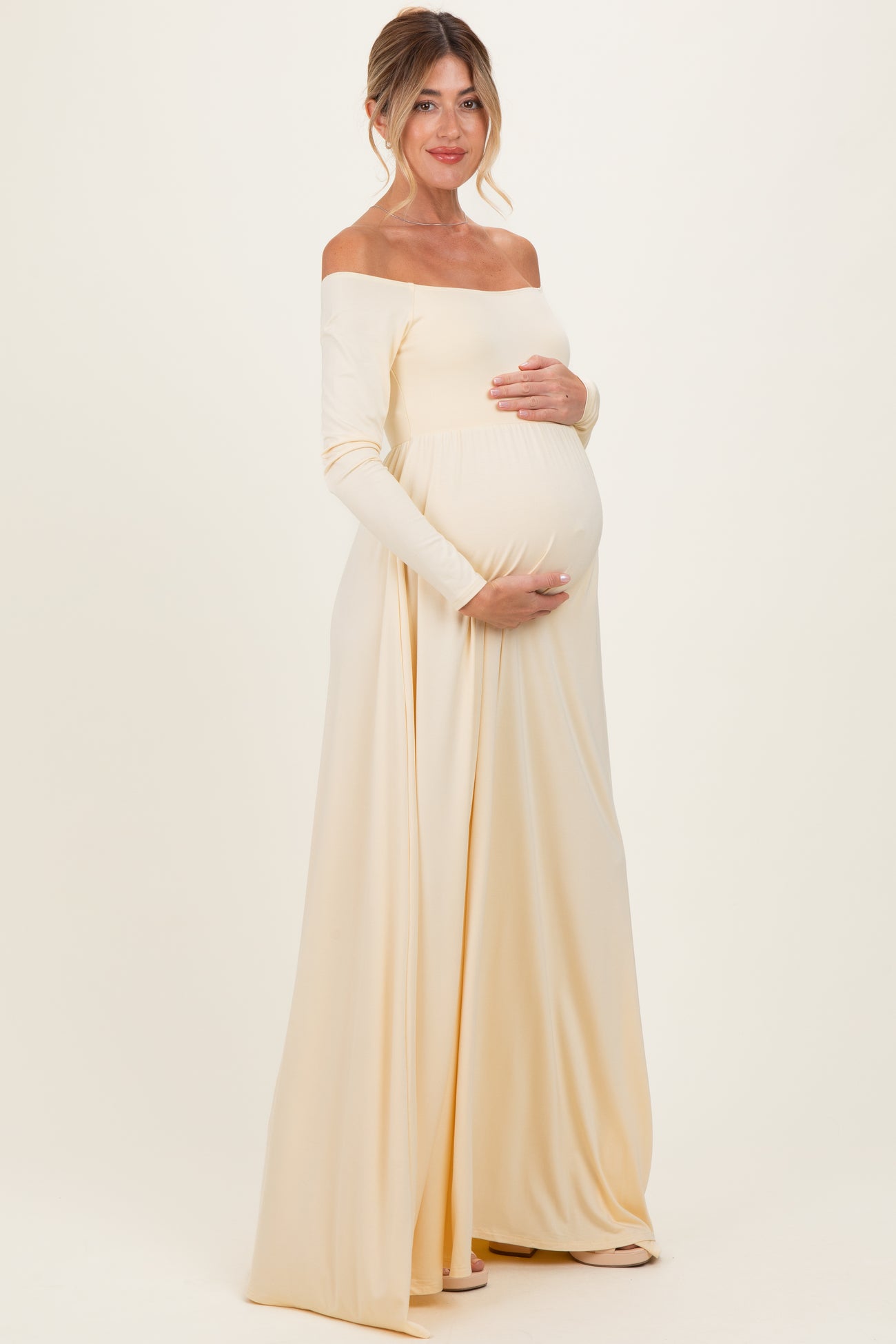 woman wearing a cream off-shoulder maternity gown, showcasing her baby bump, standing gracefully against a neutral background