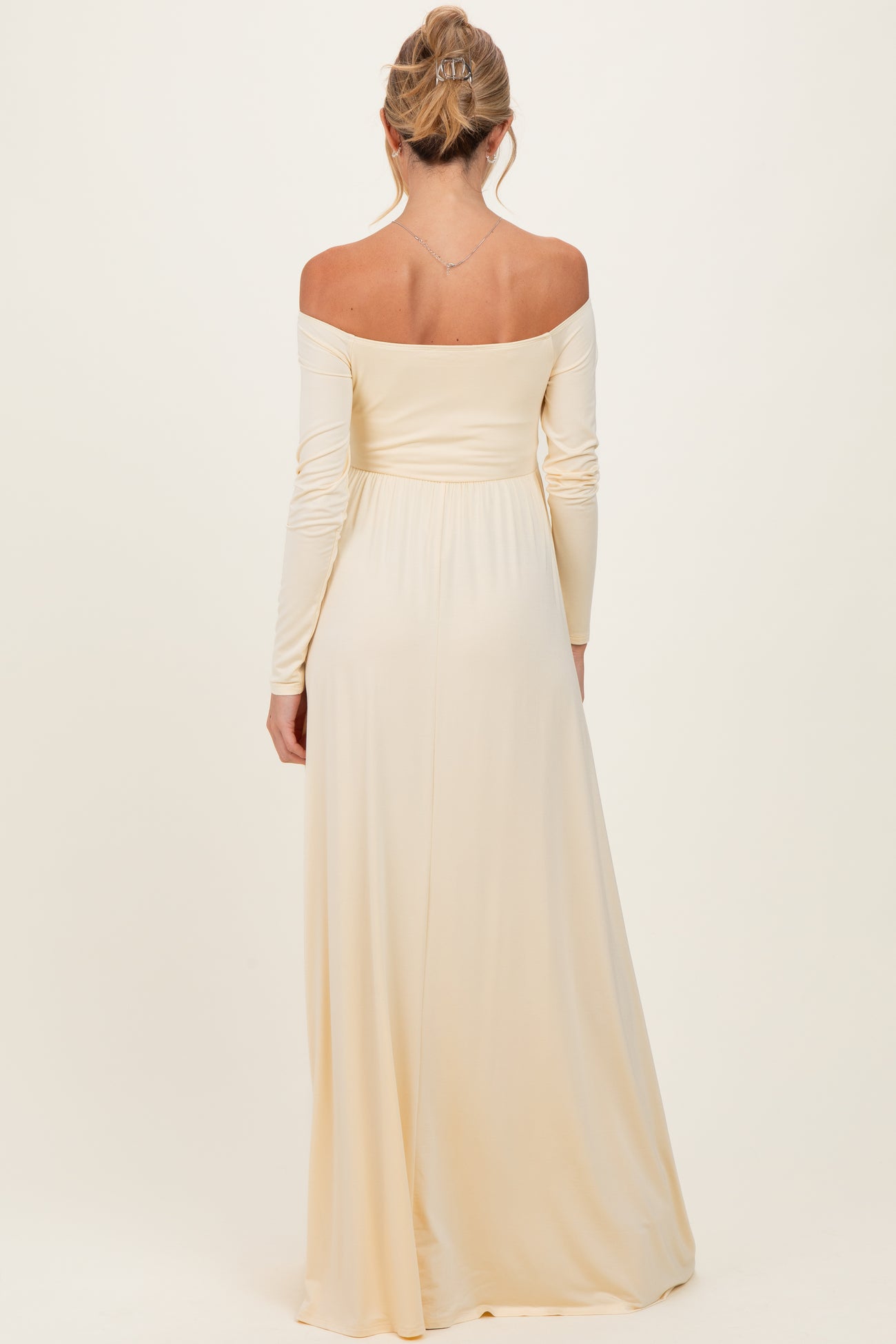 woman wearing a light beige off-the-shoulder maternity gown with long sleeves, showing a flowing silhouette from the back