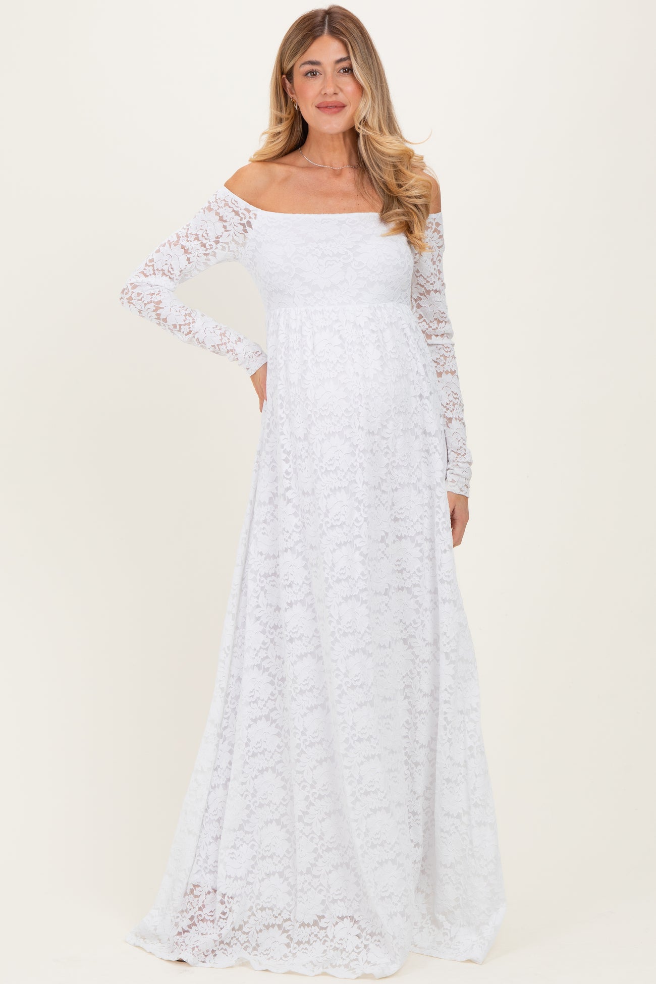 woman wearing a white lace off-shoulder maternity dress with long sleeves and a flowing skirt, showcasing her baby bump