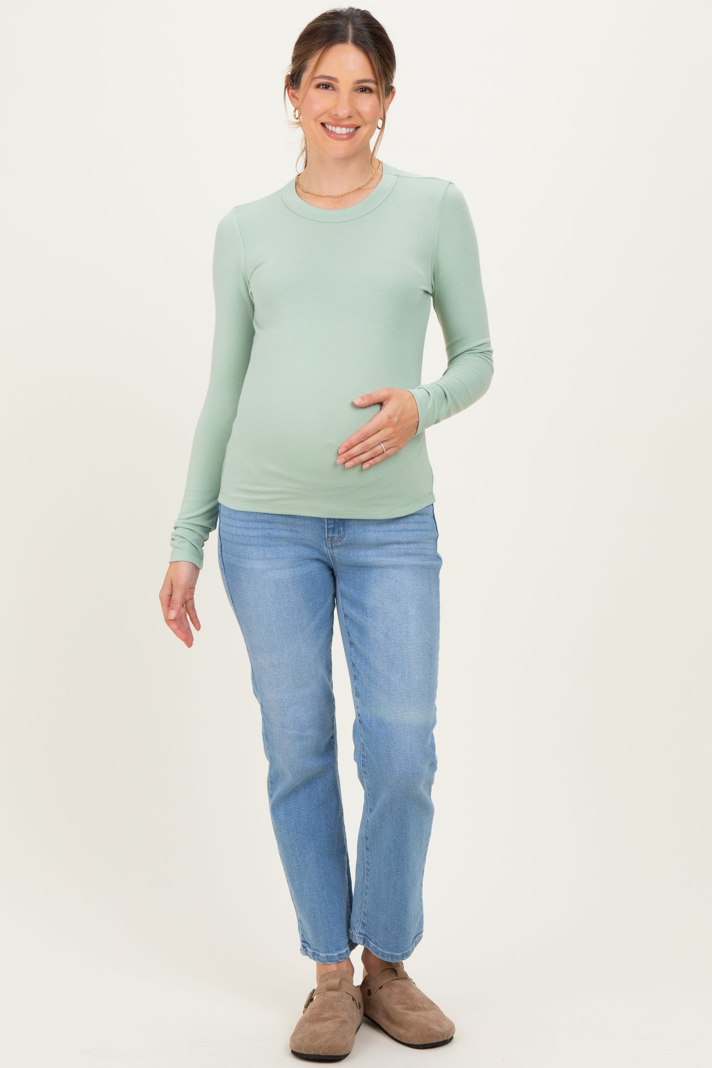 woman wearing a light green long sleeve maternity top and blue jeans, gently cradling her baby bump