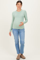 woman wearing a light green long sleeve maternity top and blue jeans, gently cradling her baby bump