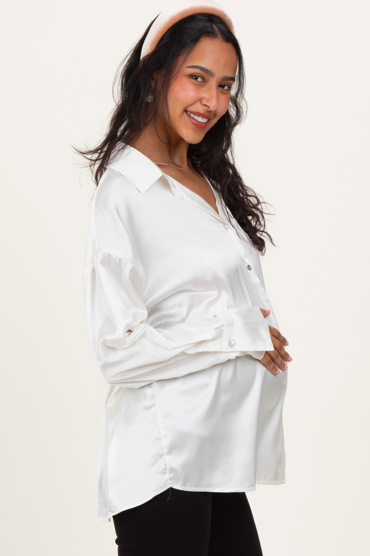 woman wearing a white satin maternity blouse and holding her baby bump, smiling with a headband on