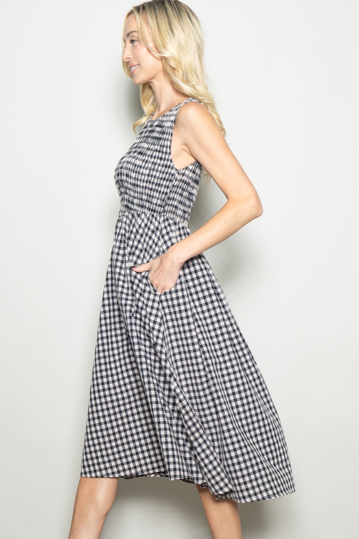 woman wearing a black and white gingham sleeveless midi dress with pockets, standing in profile