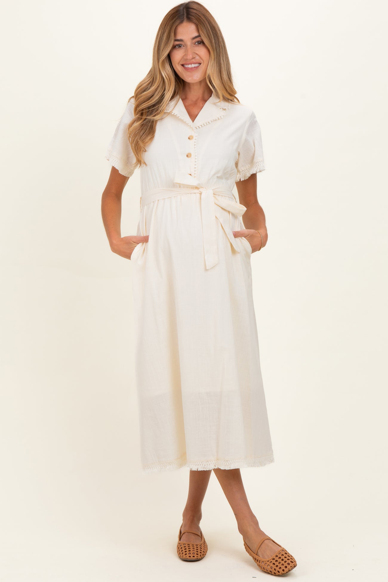 woman wearing a cream maternity dress with short sleeves, button details, and a waist tie, featuring side pockets