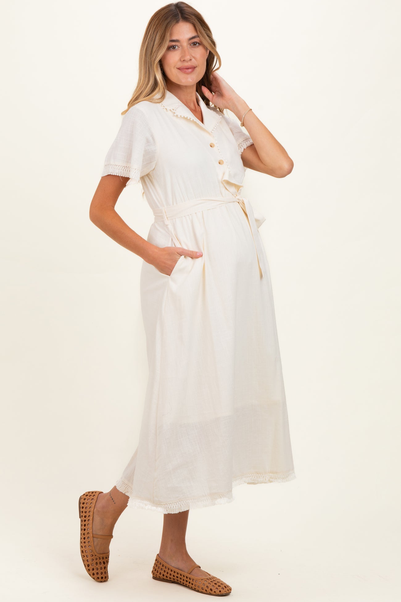 woman wearing a cream maternity dress with short sleeves, buttons, and a tie waist, featuring pockets and a visible baby bump