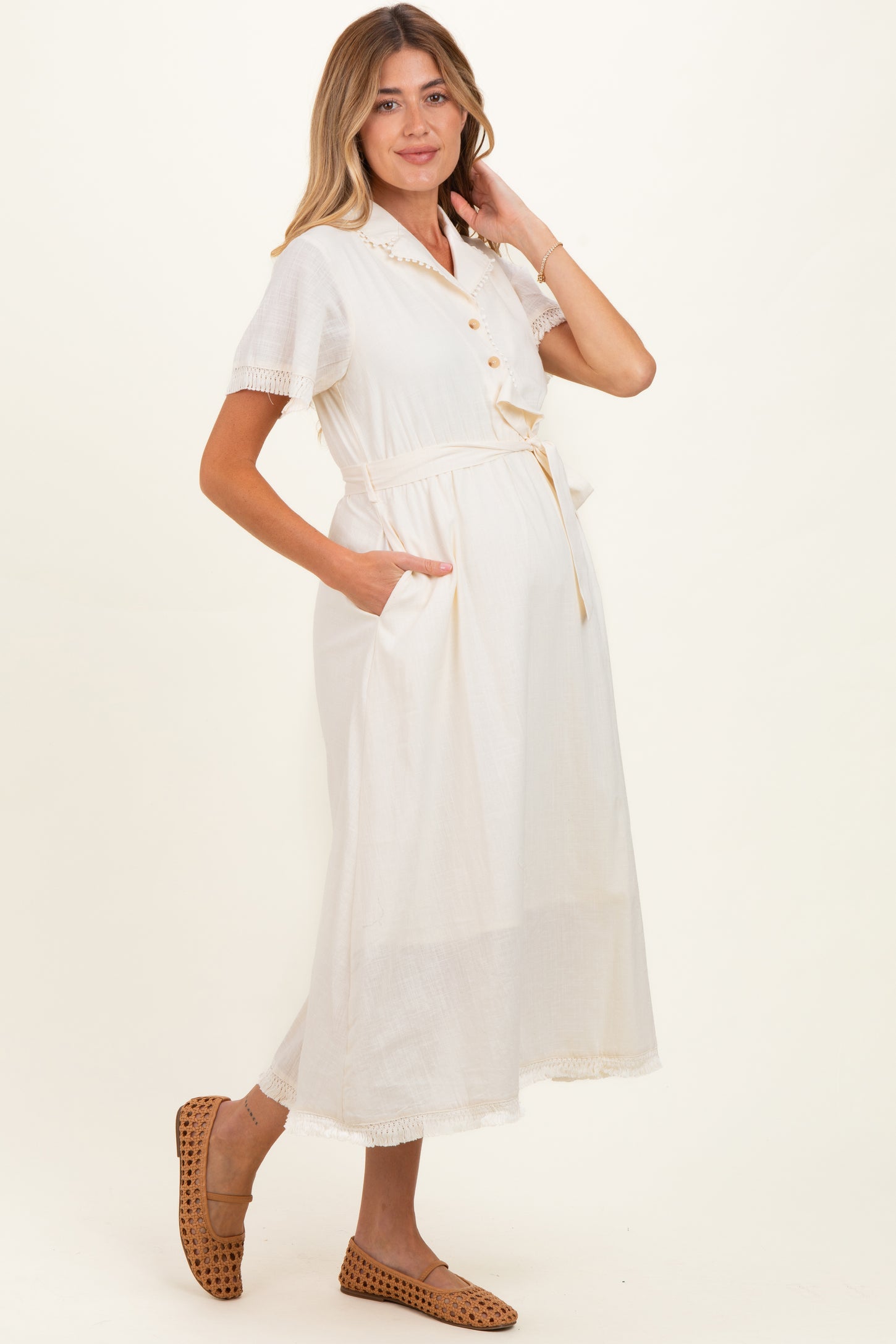 woman wearing a cream maternity dress with short sleeves, buttons, and a tie waist, featuring pockets and a visible baby bump