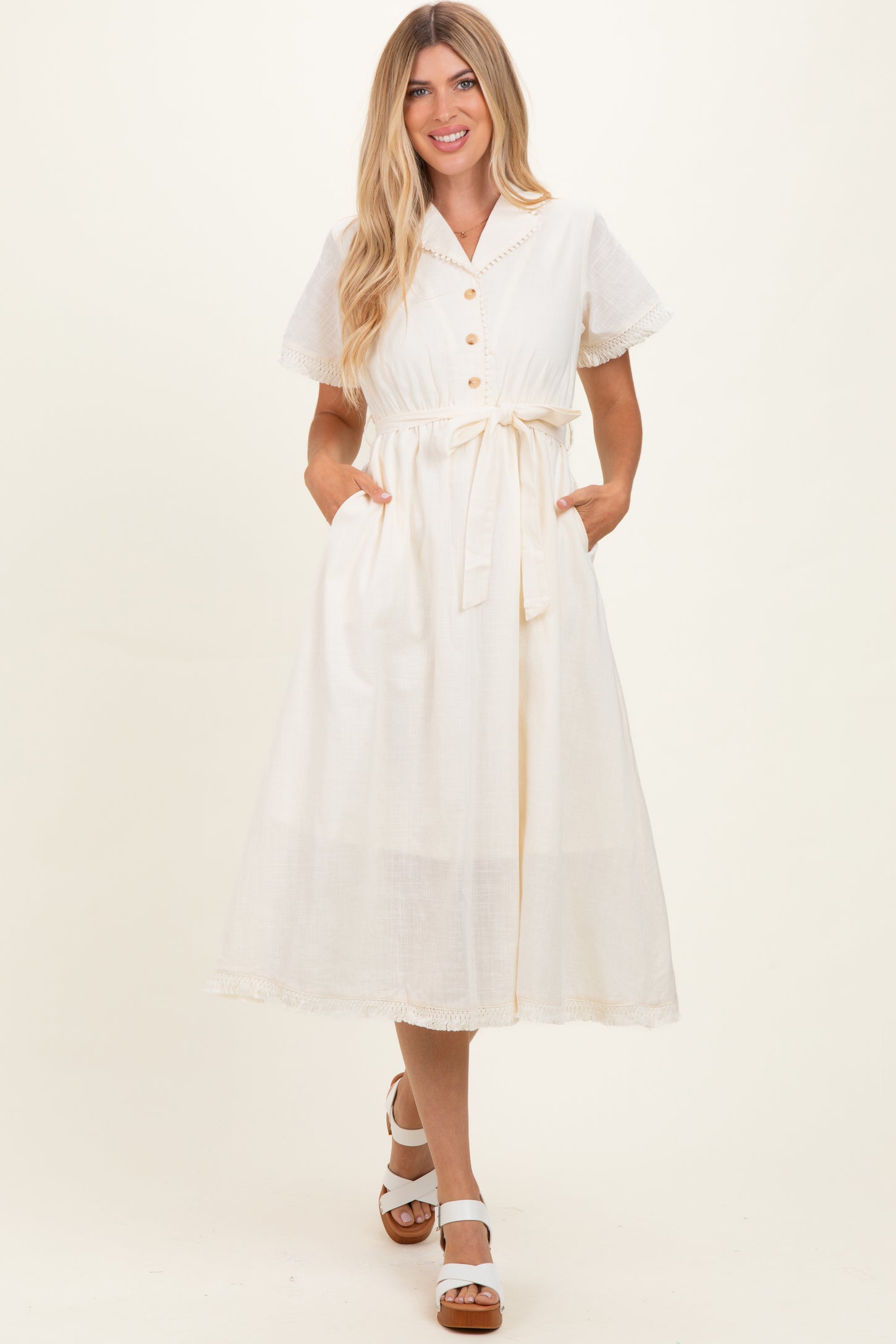 woman wearing a cream midi dress with short sleeves, button detail, tie waist, and pockets