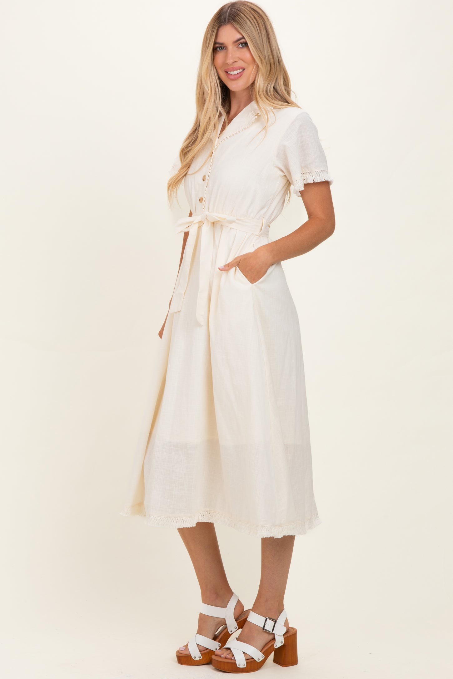 woman wearing a cream short sleeve midi dress with buttons and a tied waist, standing with hands in pockets