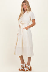 woman wearing a cream short sleeve midi dress with buttons and a tied waist, standing with hands in pockets