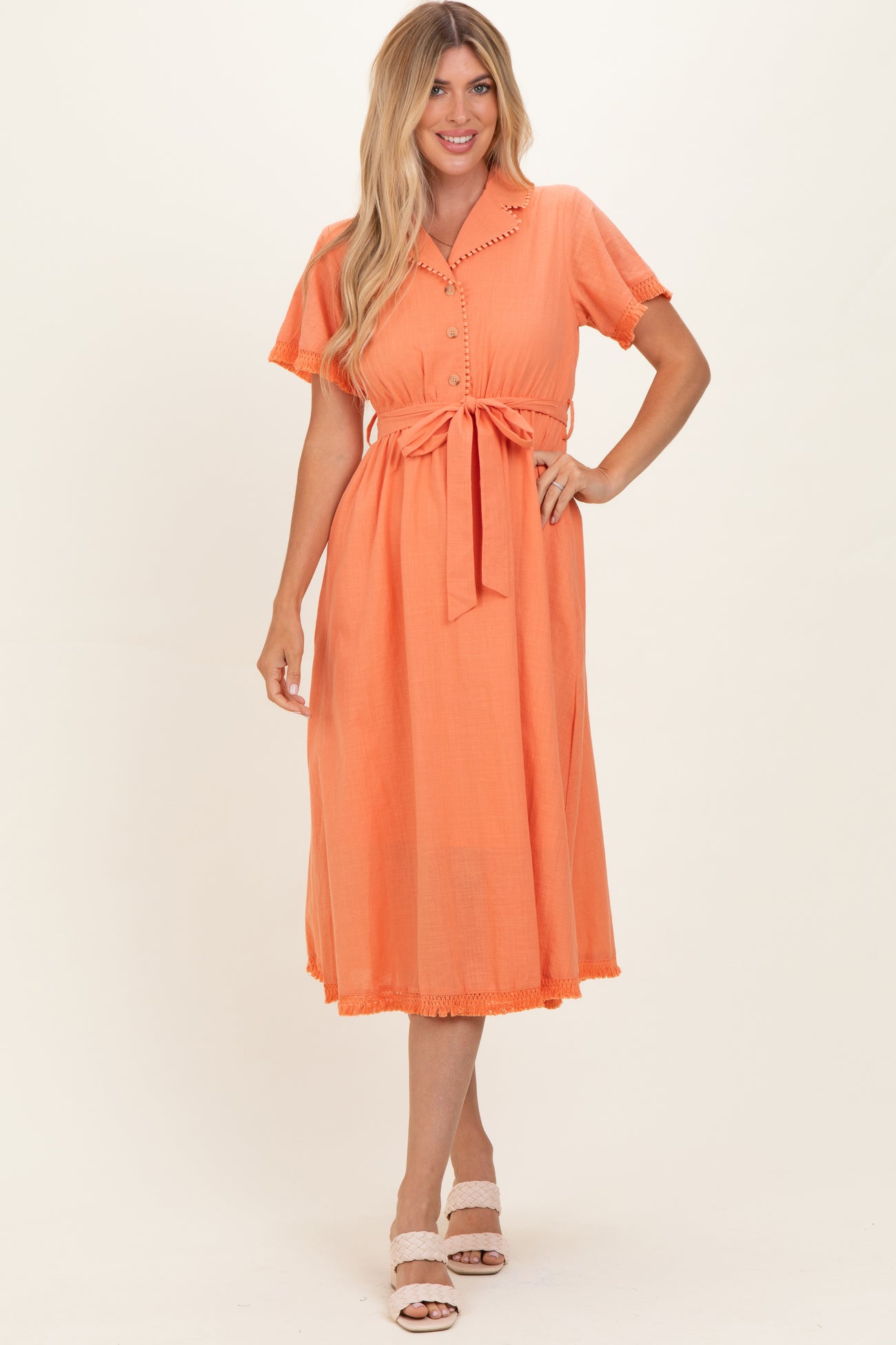 woman wearing a coral midi shirt dress with short sleeves, a tie waist, and fringe detailing at the hem
