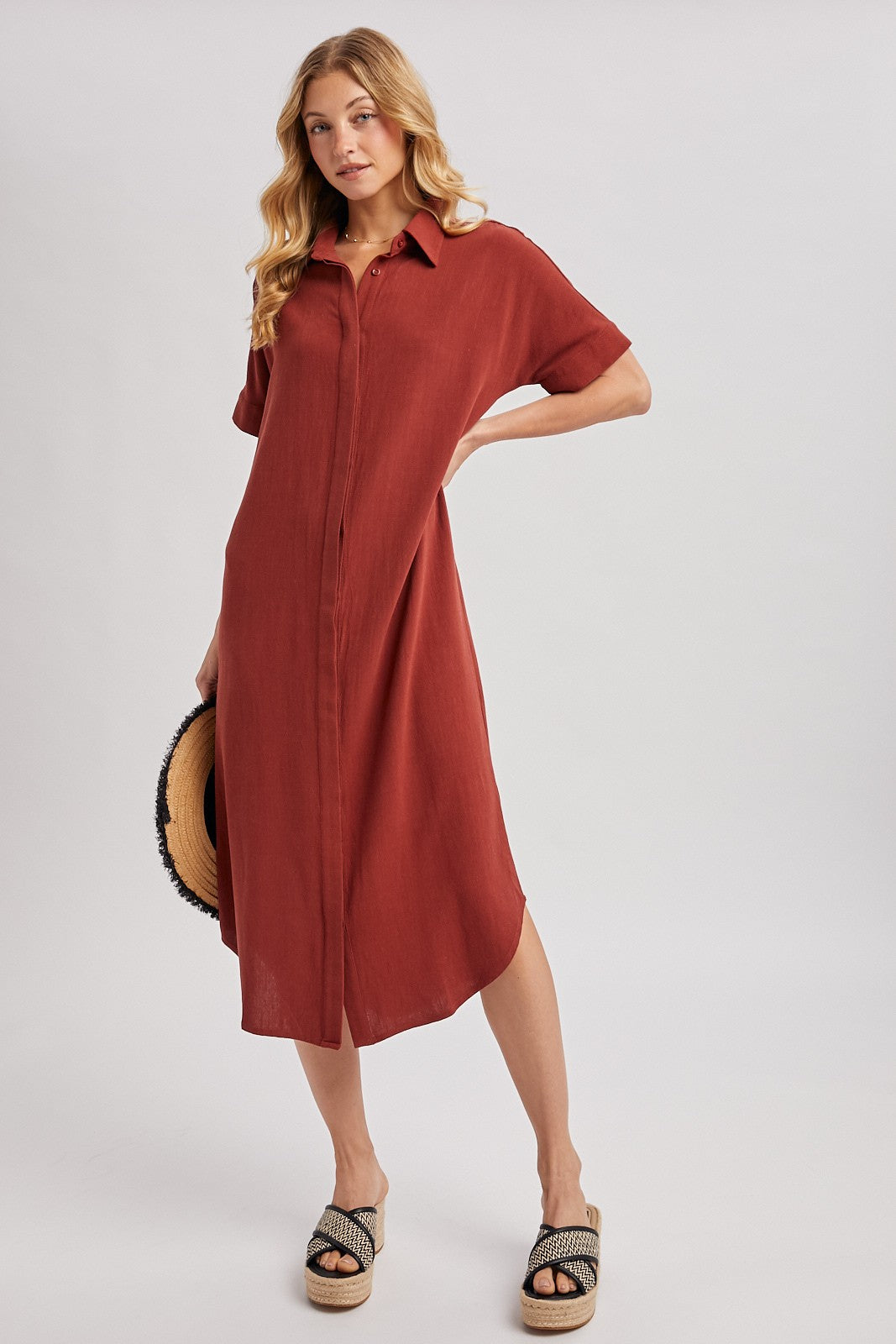woman wearing a rust-colored button-up midi dress with short sleeves and a collar, accessorized with a straw hat and platform sandals