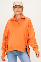woman wearing an orange oversized maternity pullover with a collared neckline and side slits, styled with a cap