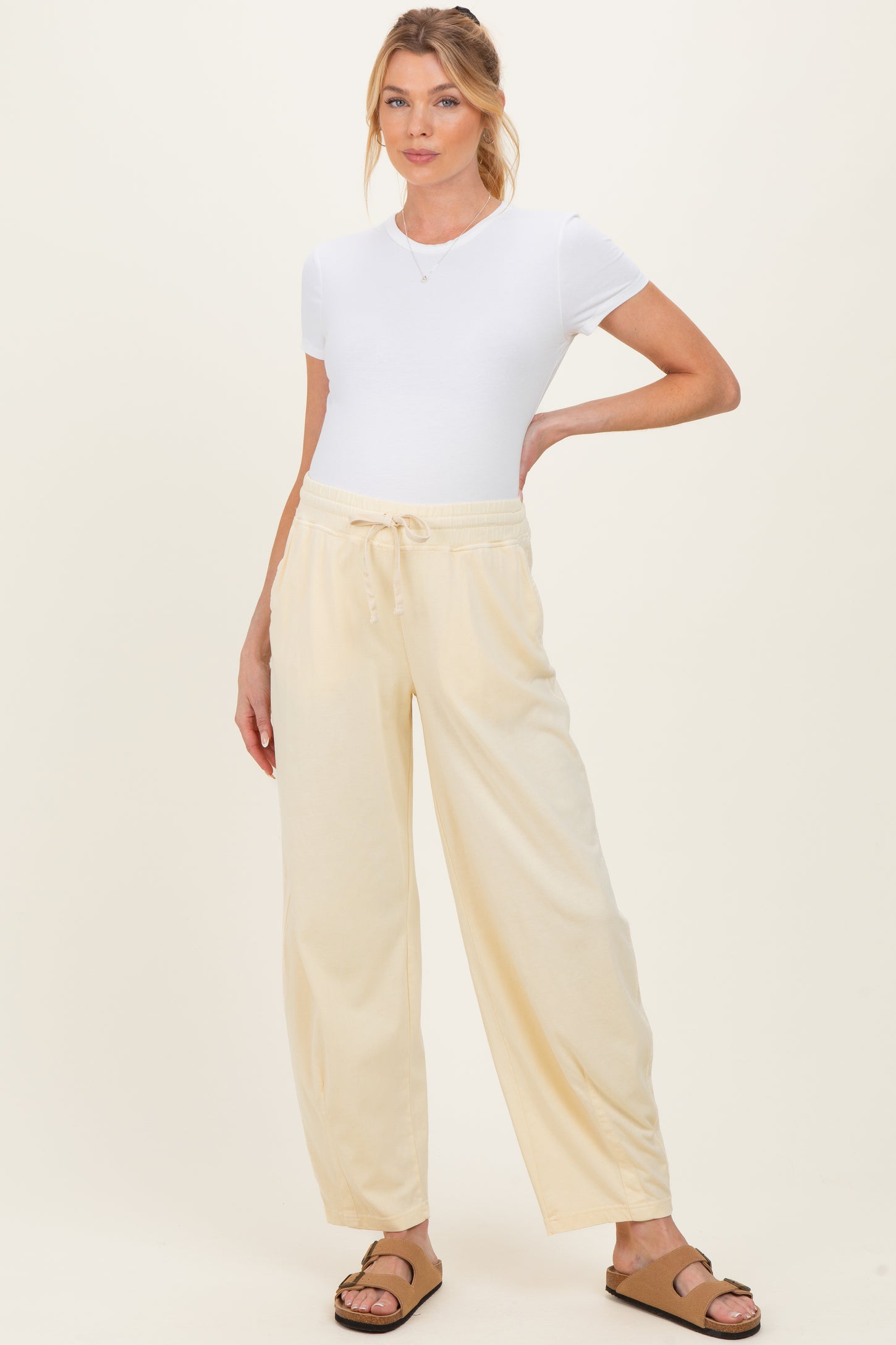 woman wearing a white short sleeve maternity top and light yellow drawstring pants, standing with one hand on her hip