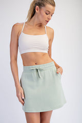 woman wearing a light green drawstring skirt and a white cropped tank top, posing with one hand on her hip