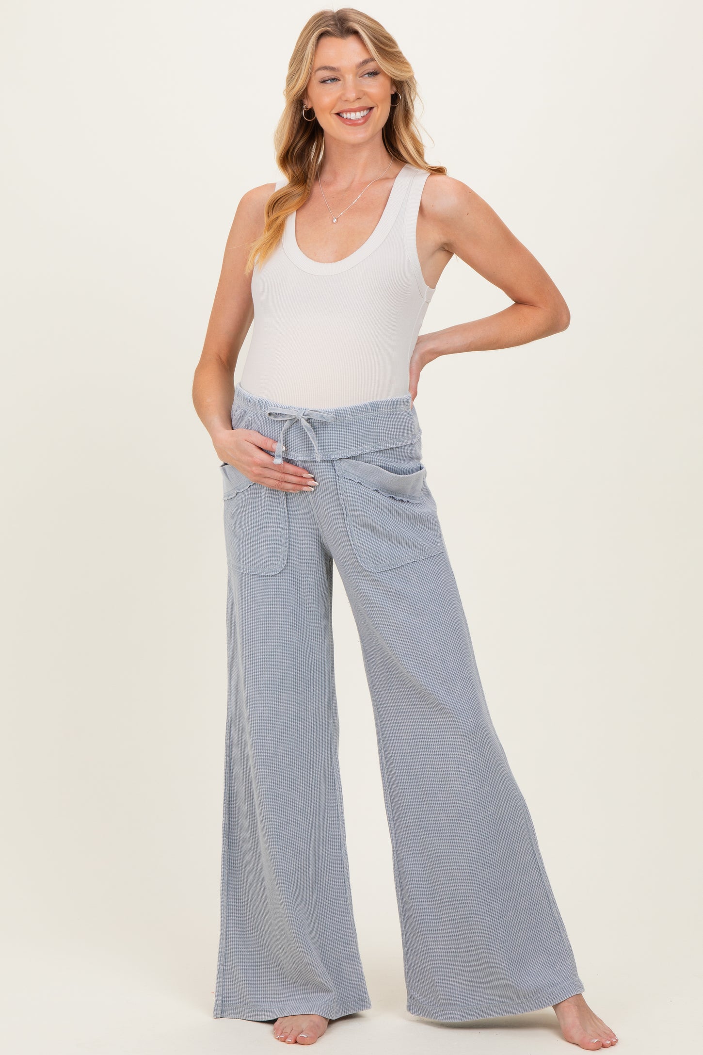 woman wearing a white tank top and light blue maternity wide leg pants, holding her baby bump