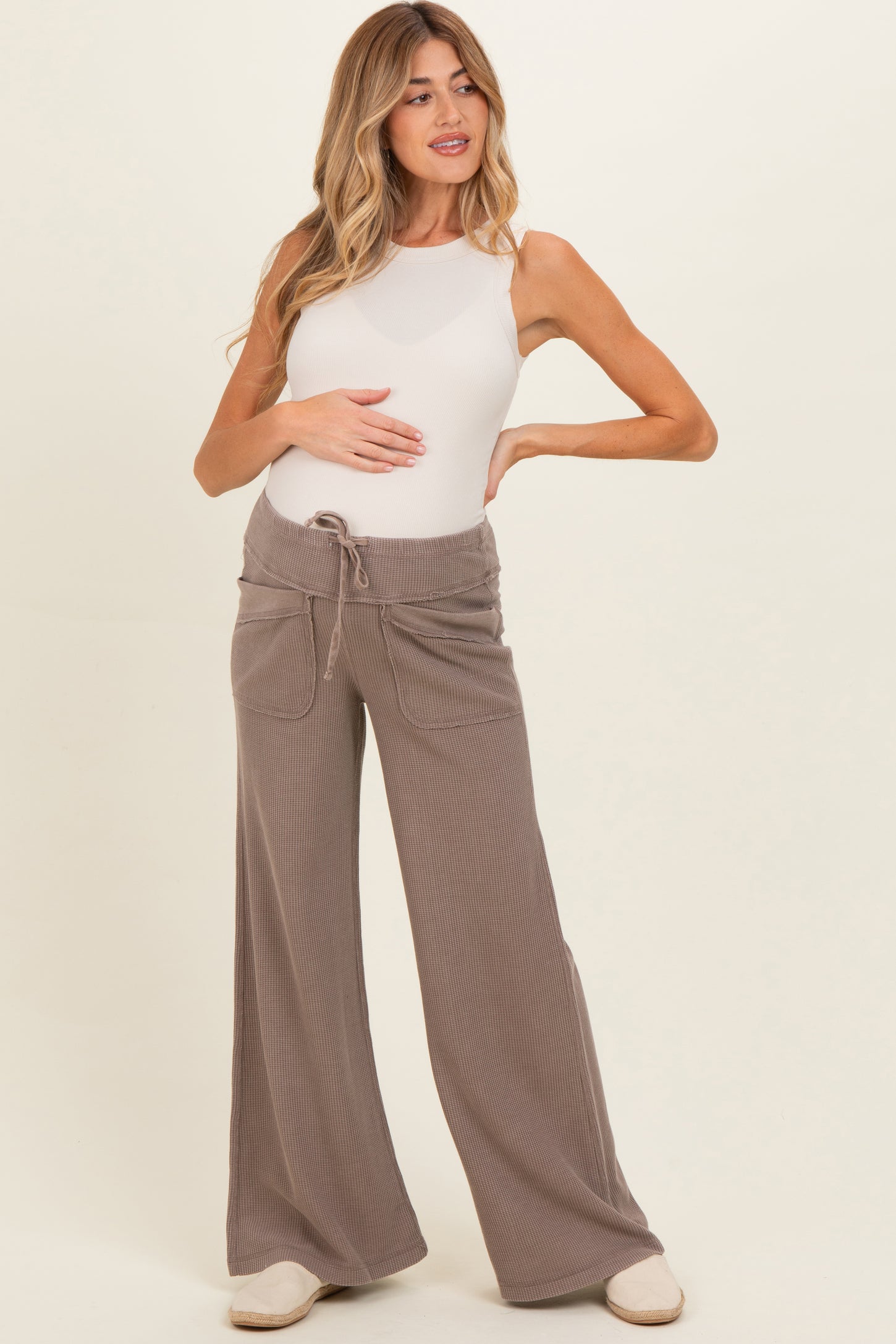 woman wearing a beige maternity tank top and wide-leg pants, holding her baby bump