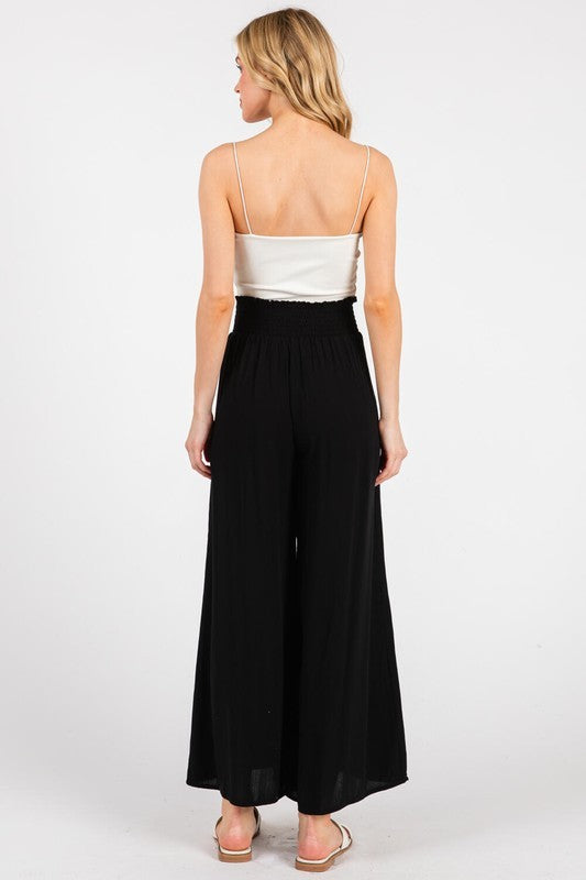 woman wearing a black wide-leg jumpsuit with a smocked waist and a satin white top, viewed from the back