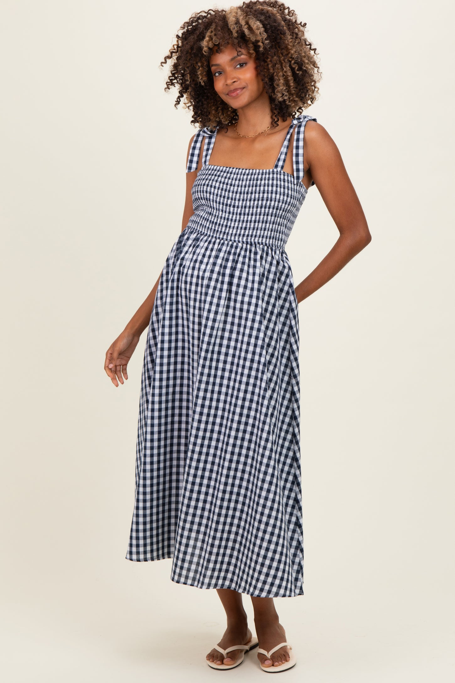 woman wearing a navy and white gingham maternity dress with smocked bodice and tie straps, showing her baby bump