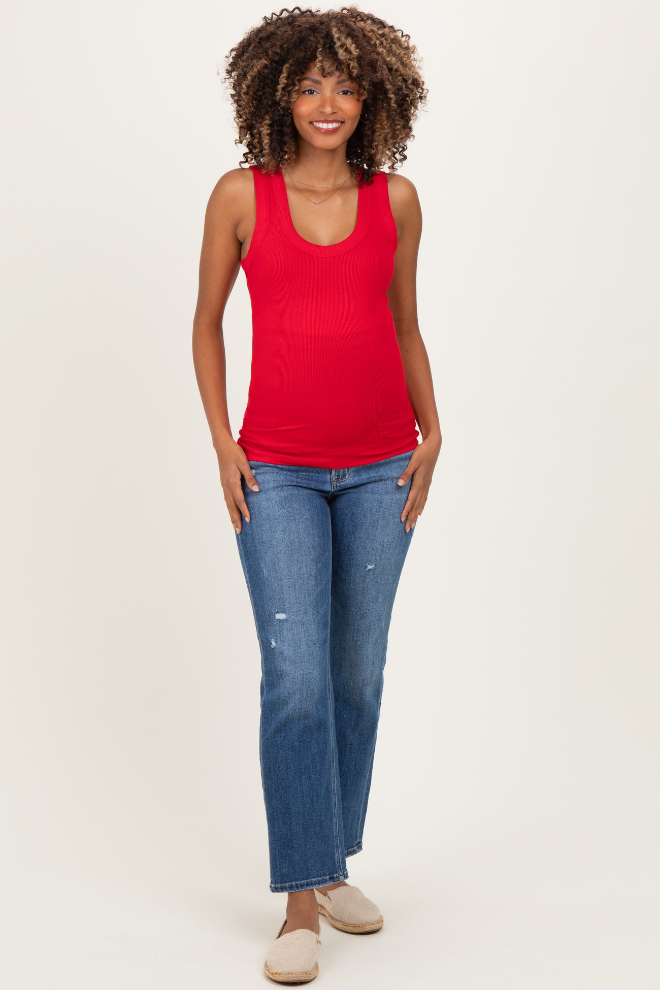 woman wearing a red maternity tank top and blue distressed jeans, showing her baby bump