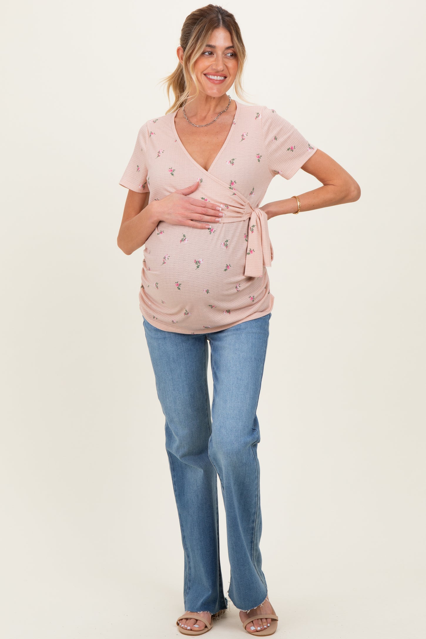 woman wearing a light pink maternity top with floral prints and a tie detail, paired with blue flared jeans and showing her baby bump