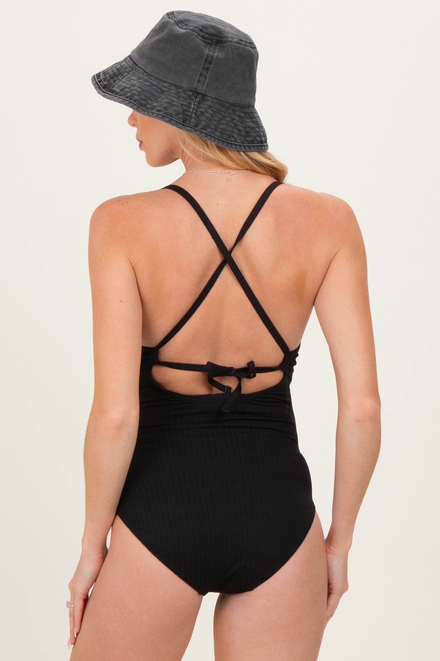 woman wearing a black ribbed maternity swimsuit with crisscross straps and a tied back, accessorized with a bucket hat