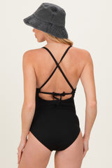 woman wearing a black ribbed maternity swimsuit with crisscross straps and a tied back, accessorized with a bucket hat