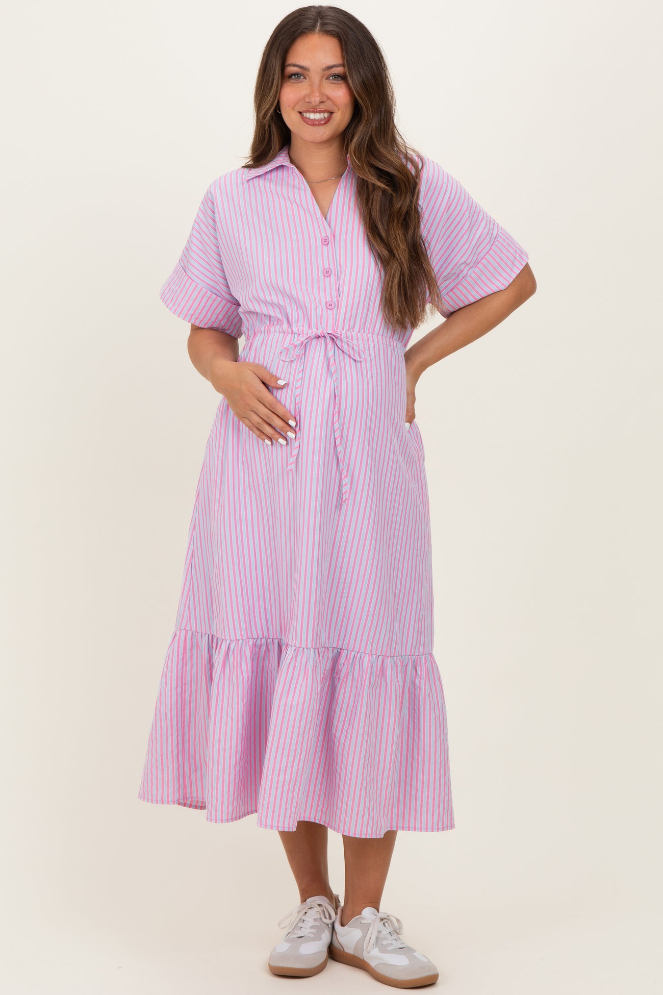 woman wearing a pink and white striped maternity dress with a tie waist and ruffled hem, gently cradling her baby bump