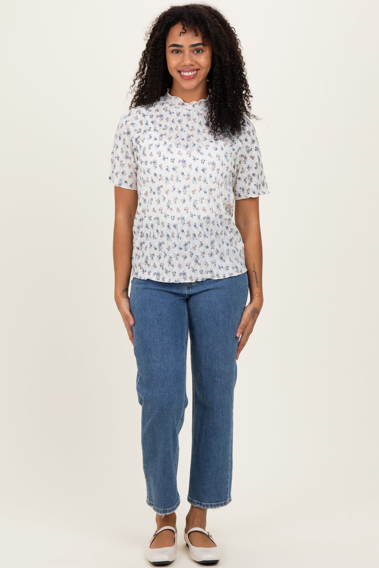 woman wearing a white short sleeve floral print top and blue high-waisted jeans standing against a neutral background