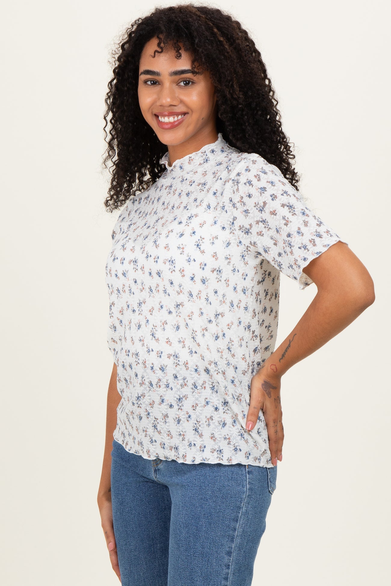 woman wearing a white short sleeve floral blouse and blue jeans, smiling at the camera