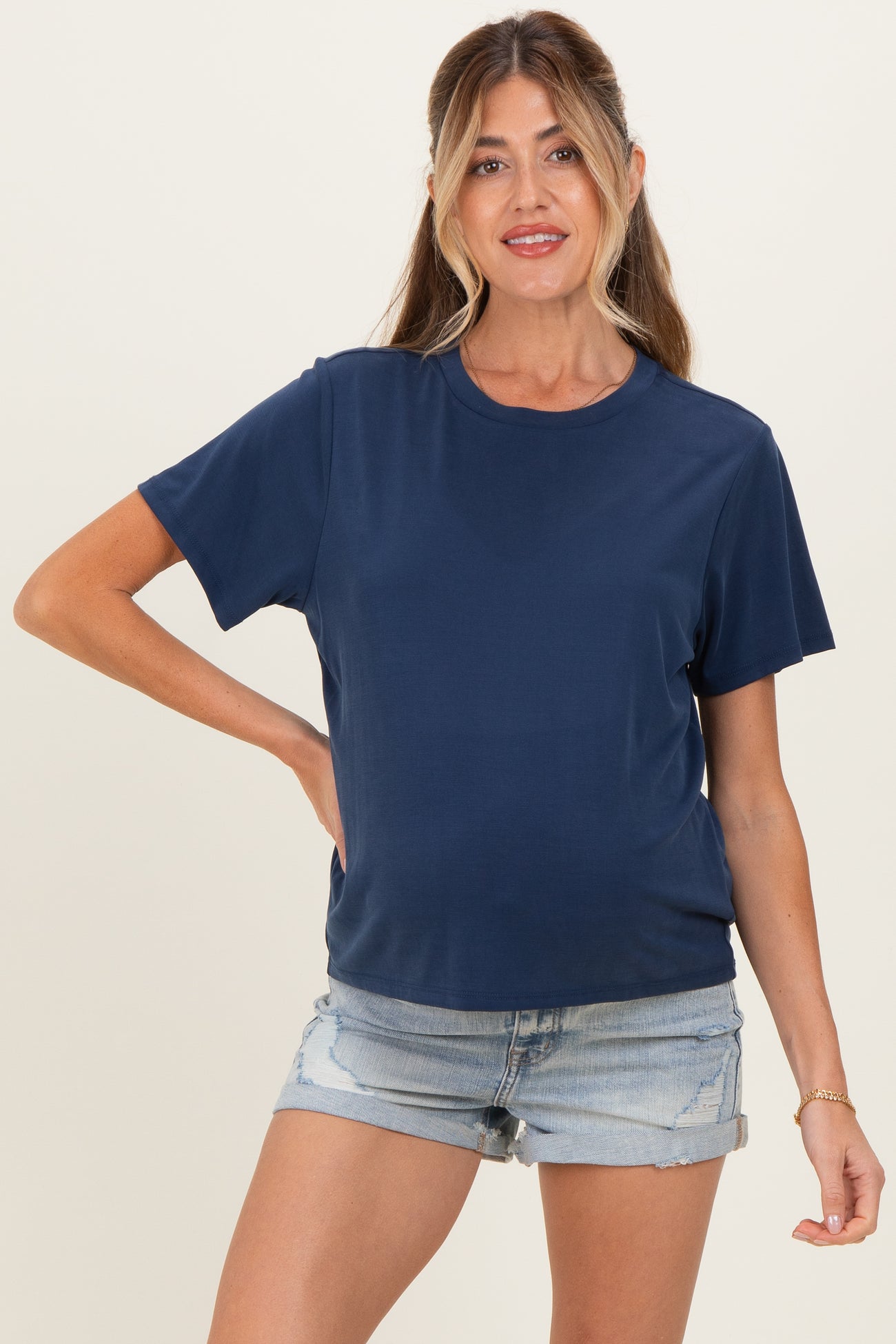 woman wearing a navy blue short sleeve maternity top and denim shorts, posing with one hand on her hip