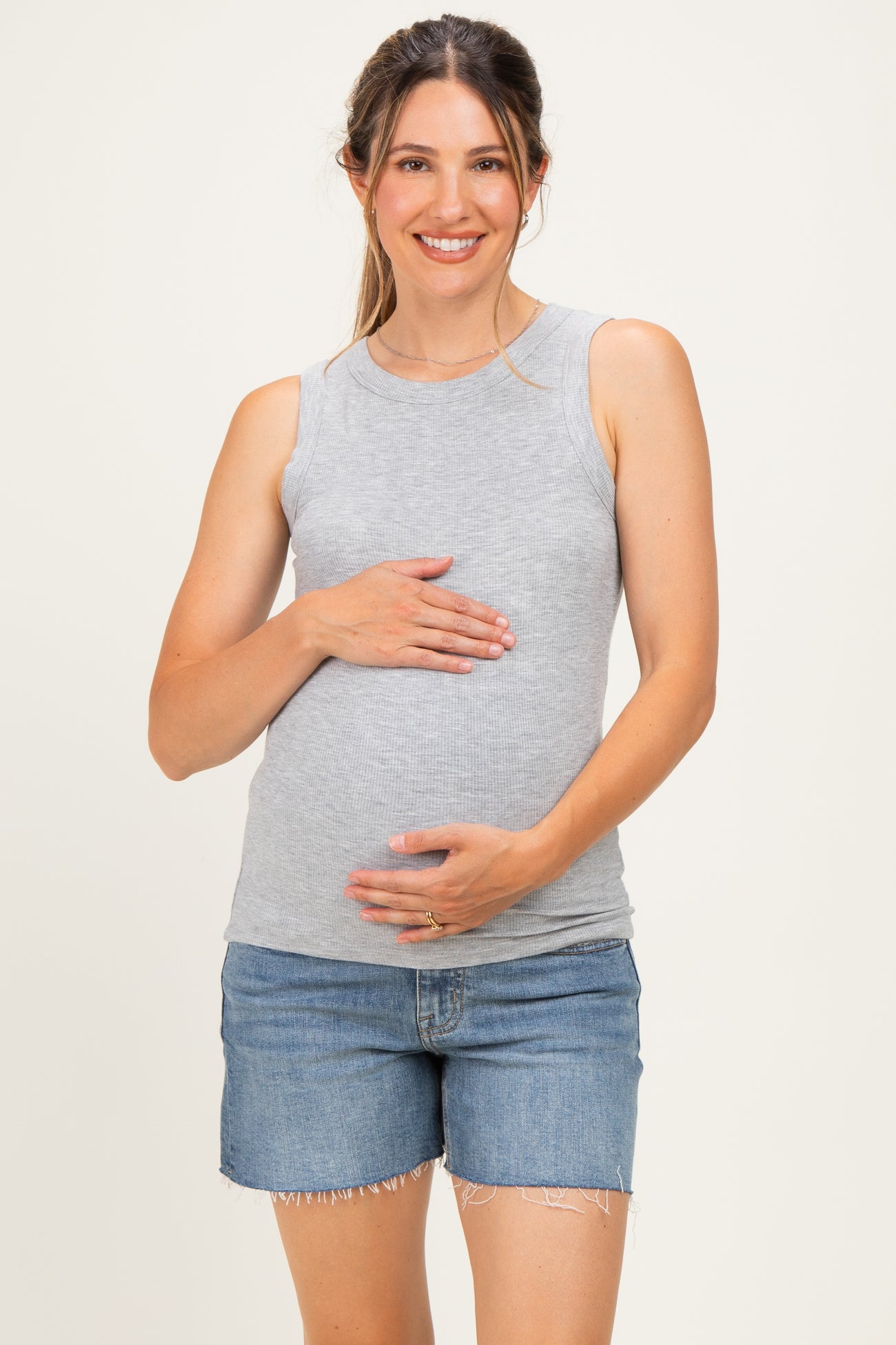 woman wearing a light gray maternity tank top and holding her baby bump while smiling, paired with denim shorts