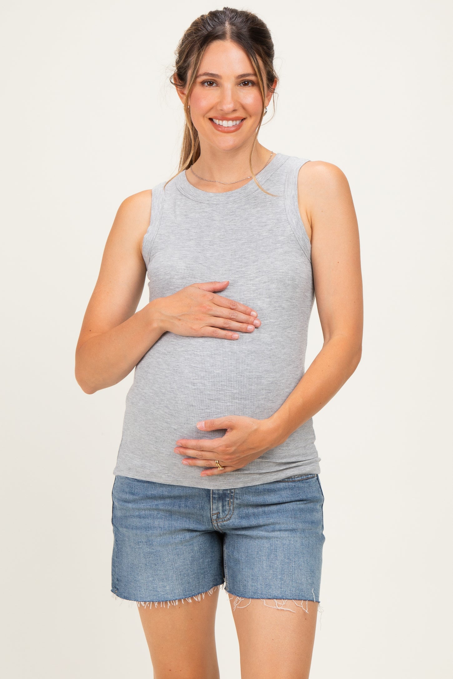 woman wearing a light gray maternity tank top and holding her baby bump while smiling, paired with denim shorts