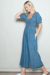 woman wearing a denim jumpsuit with a smocked bodice and puff sleeves, hands in pockets, posing sideways