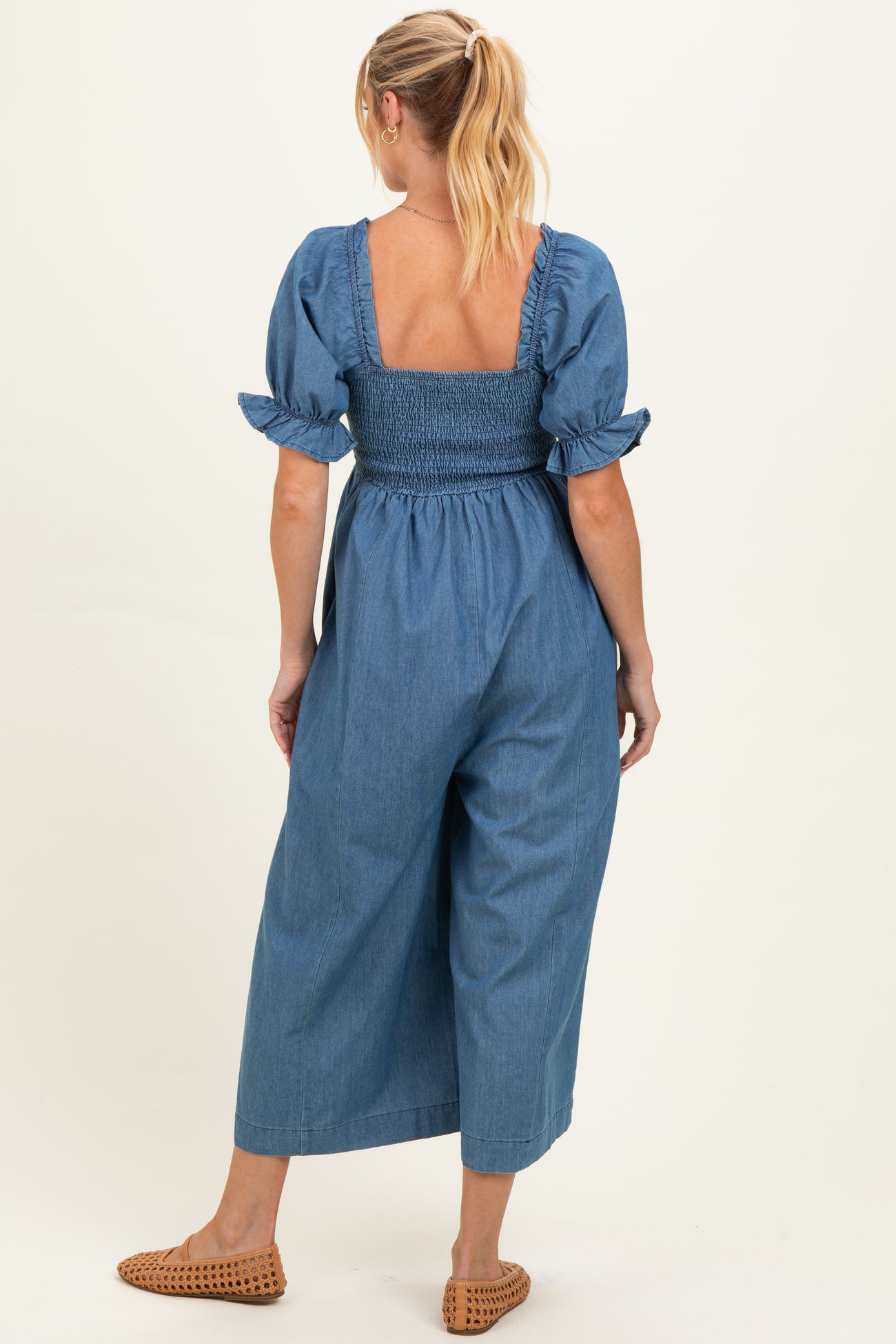 woman wearing a denim maternity jumpsuit with a smocked bodice and puff sleeves, showing her baby bump from the back