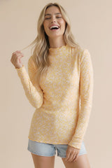 woman wearing a yellow long sleeve top with a floral pattern and a scalloped neckline, smiling against a beige background