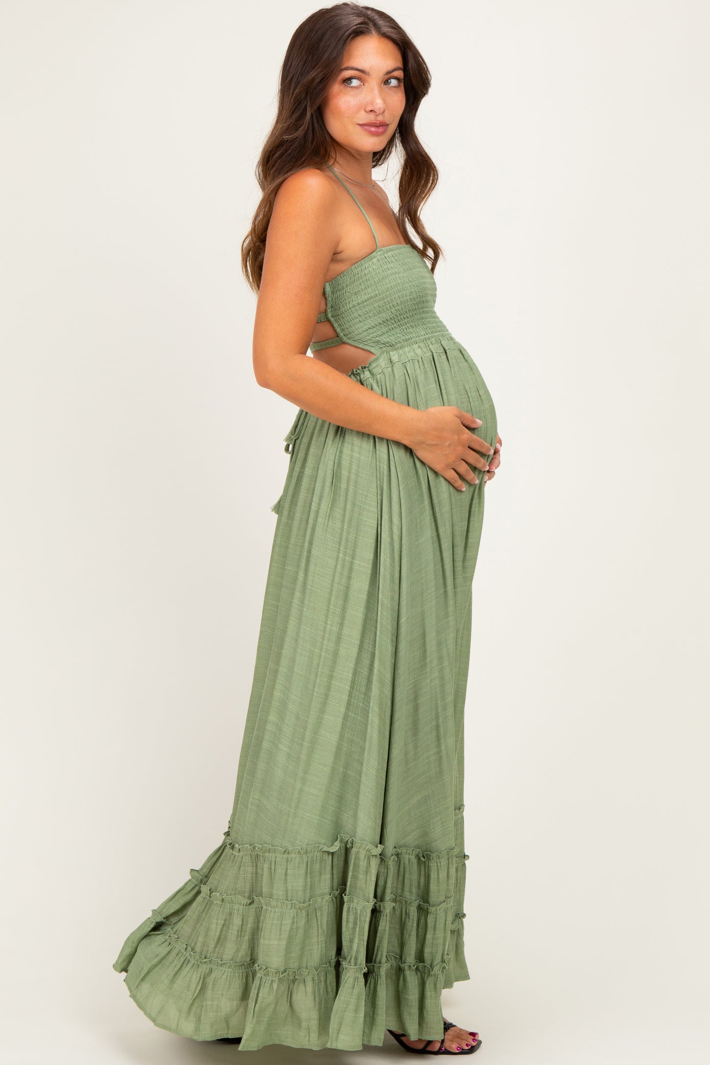 woman wearing a green maternity maxi dress with a smocked bodice and ruffle hem, gently cradling her baby bump