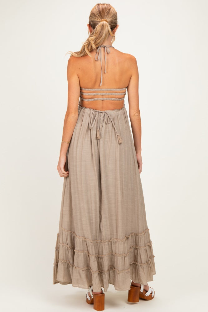 woman wearing a beige maternity maxi dress with a ruffled hem and an open back, showing ties at the neck and waist