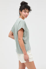 woman wearing a light green short sleeve top and cream-colored shorts, posing with her back turned to the camera