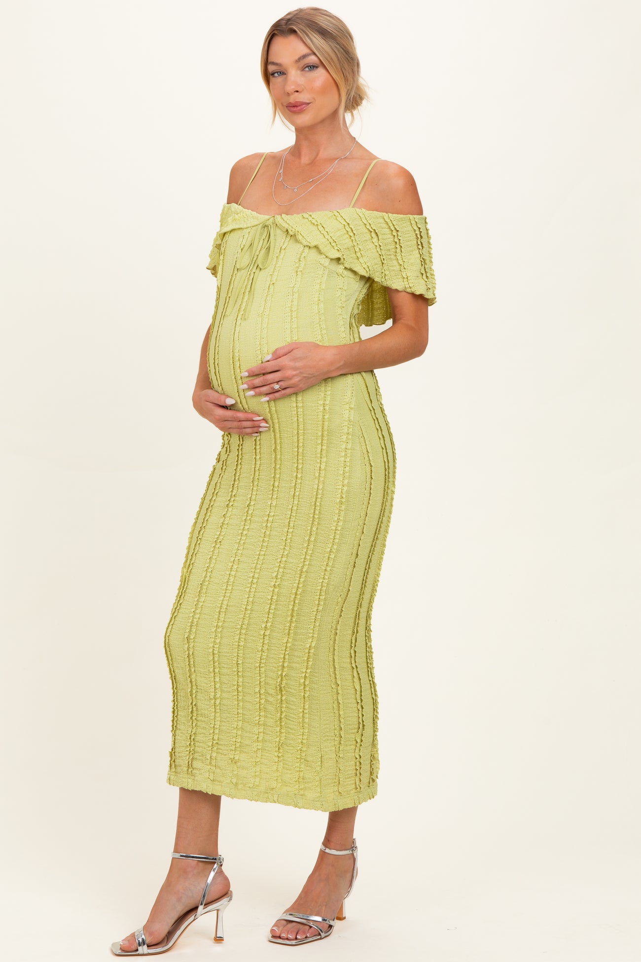 woman wearing a light green off-the-shoulder maternity dress with a tie detail, gently cradling her baby bump