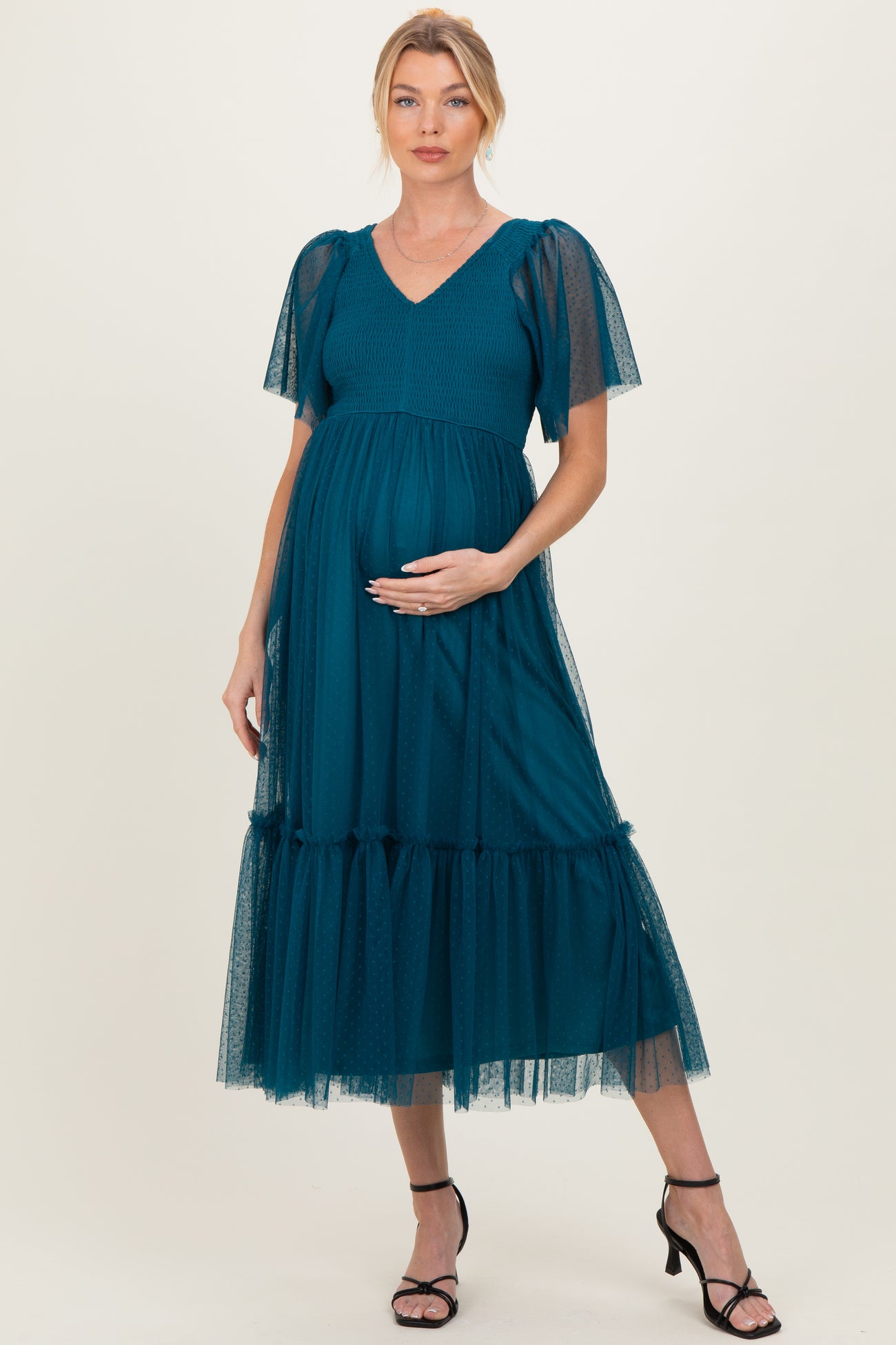 woman wearing a teal maternity dress with sheer puff sleeves and a tiered tulle skirt, holding her baby bump
