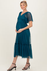woman wearing a teal maternity dress with short sheer sleeves and a tiered tulle skirt, holding her baby bump