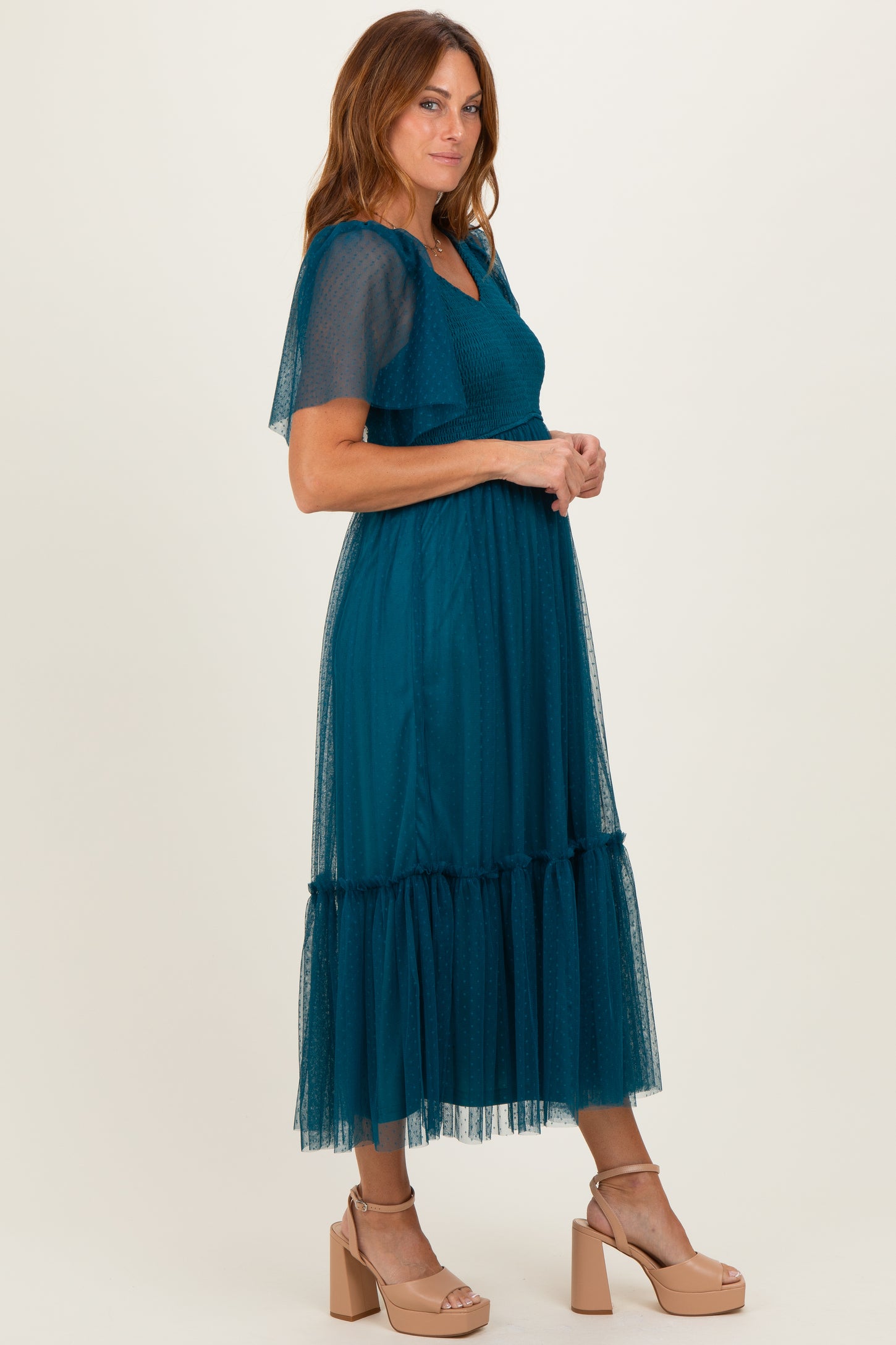 woman wearing a teal tulle midi dress with flutter sleeves and a ruffled hem, posing sideways against a neutral background