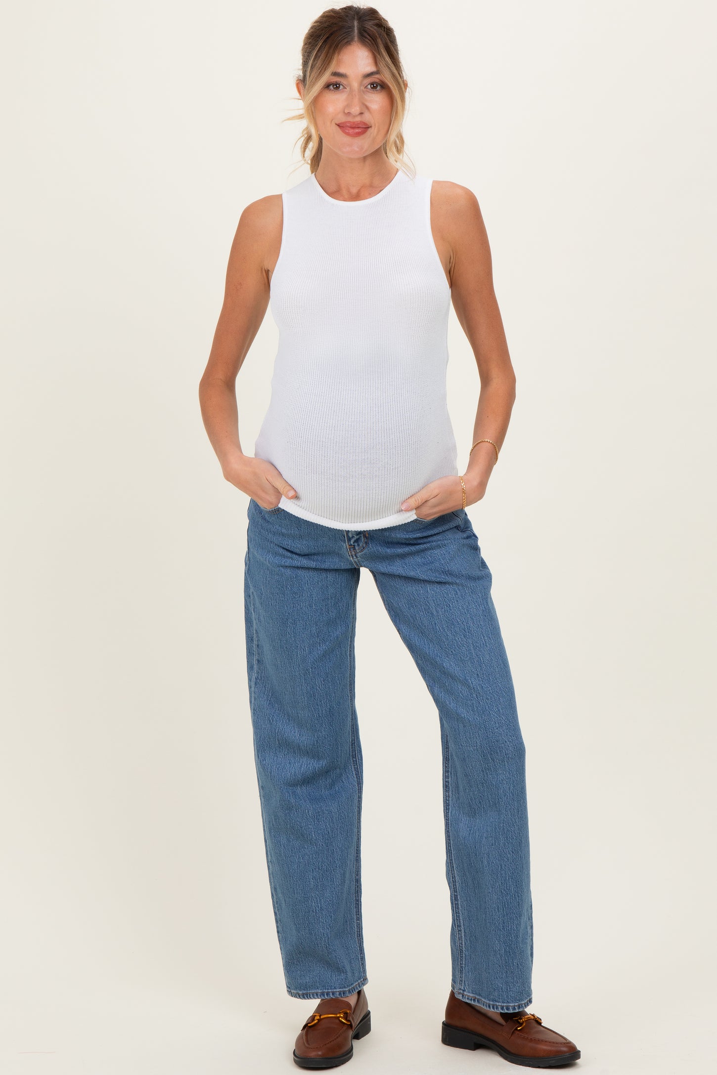 woman wearing a white sleeveless maternity top and blue wide-leg jeans, showing a visible baby bump