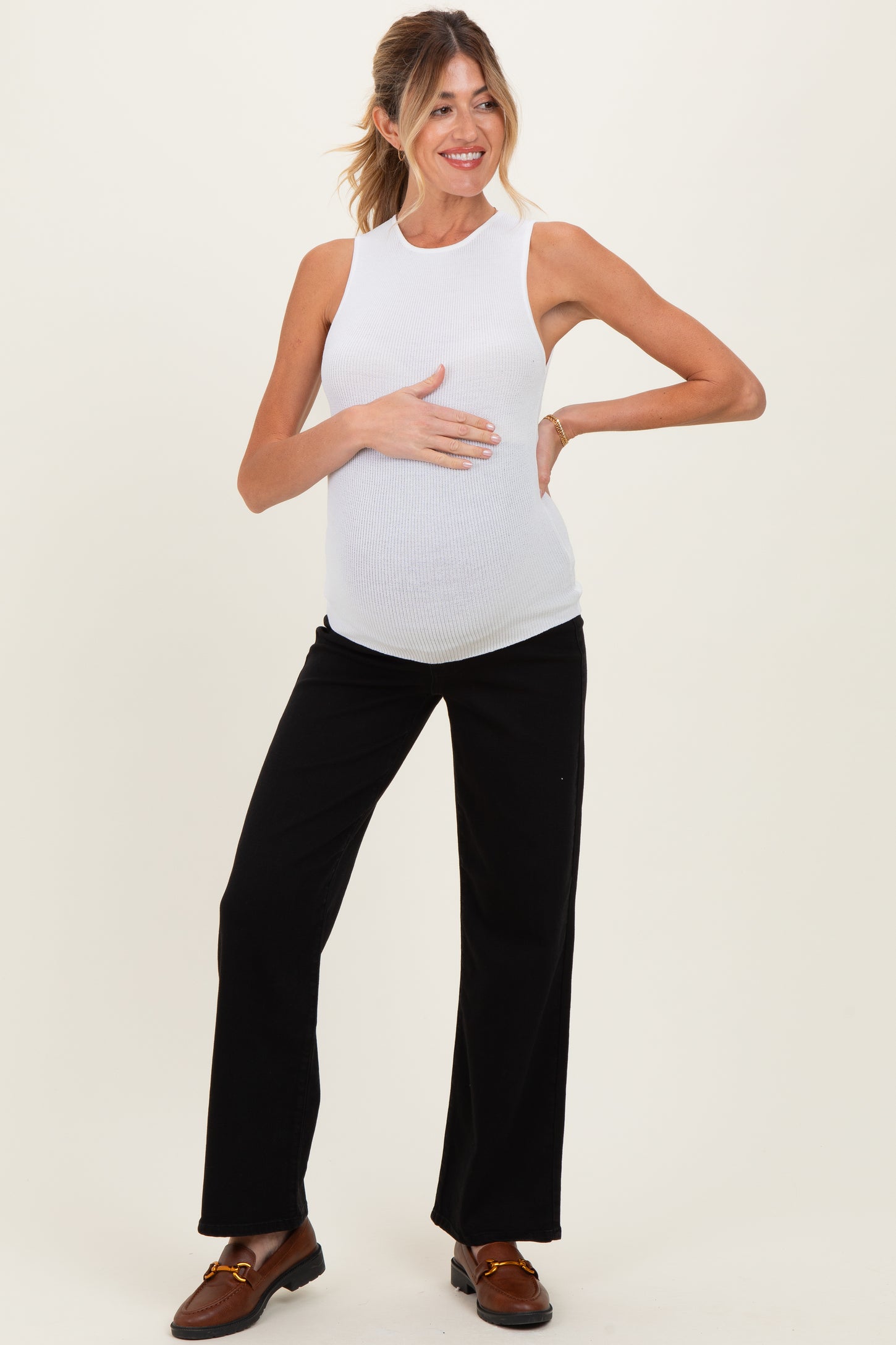 woman wearing a white sleeveless maternity top and black wide-leg pants, smiling and holding her baby bump