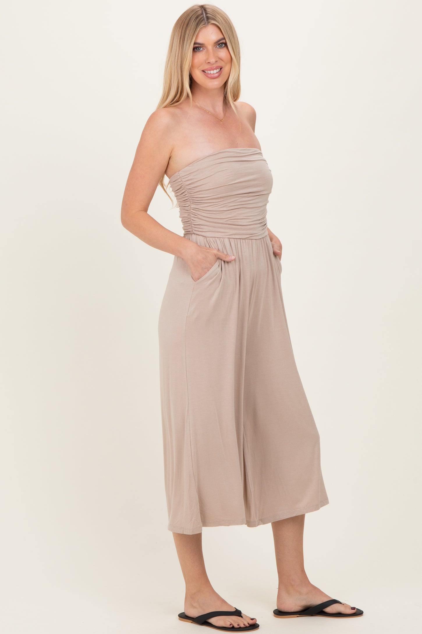 woman wearing a beige strapless jumpsuit with a ruching detail and pockets, styled with black sandals