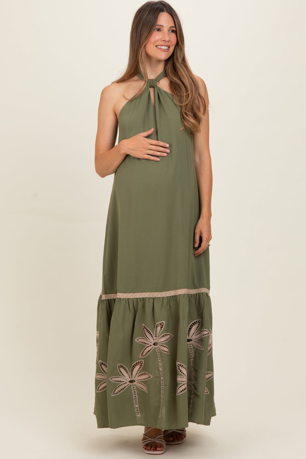 woman wearing an olive green maternity maxi dress with a tie neck and embroidered palm details, holding her baby bump