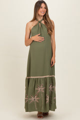 woman wearing an olive green maternity maxi dress with a tie neck and embroidered palm details, holding her baby bump