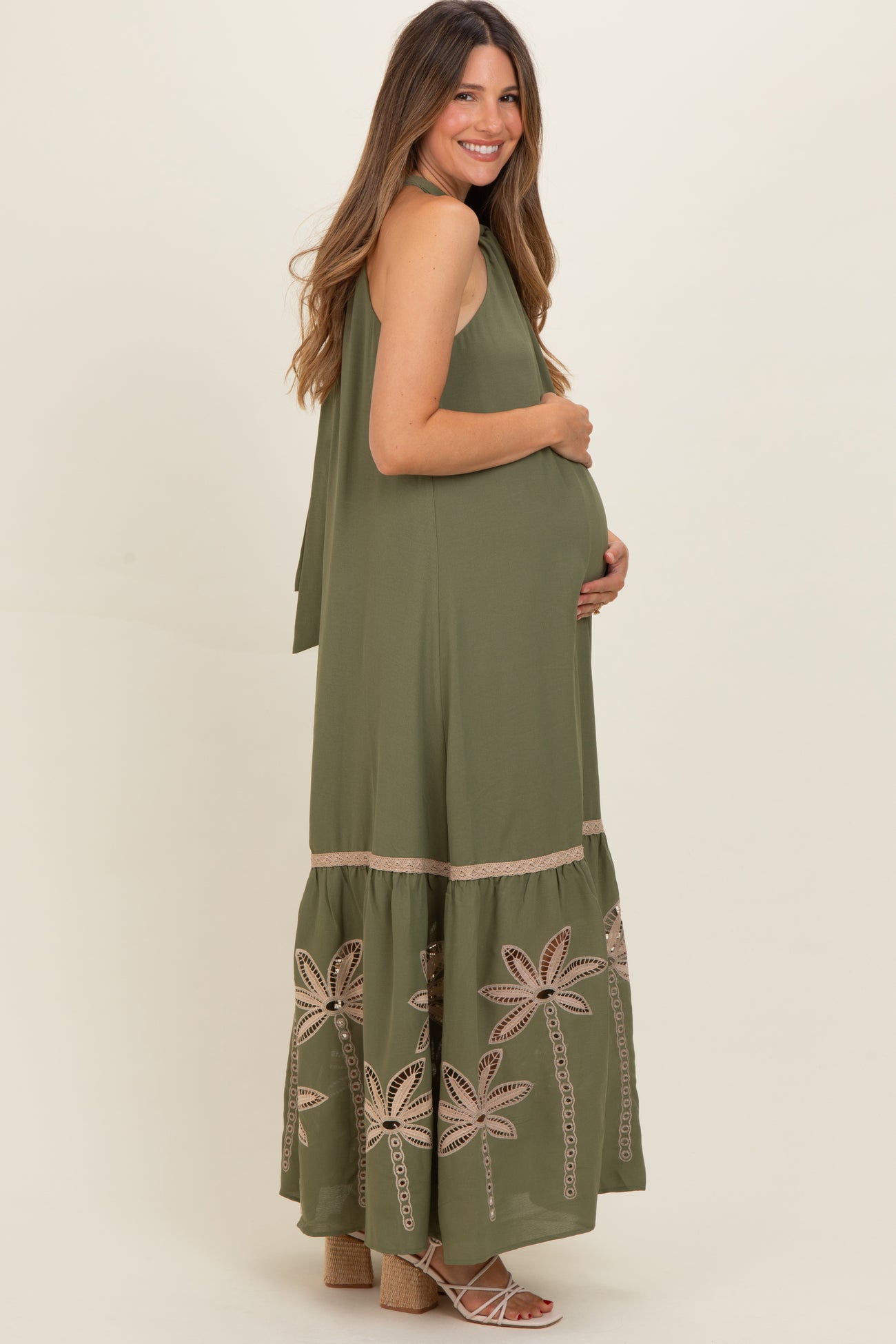 woman wearing an olive green sleeveless maternity maxi dress with floral embroidery and holding her baby bump